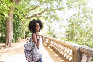 A Black woman stands on a bridge in a park holding a backpack