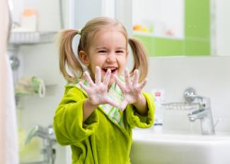 A little girl in a green bathrobe holds up soapy hands